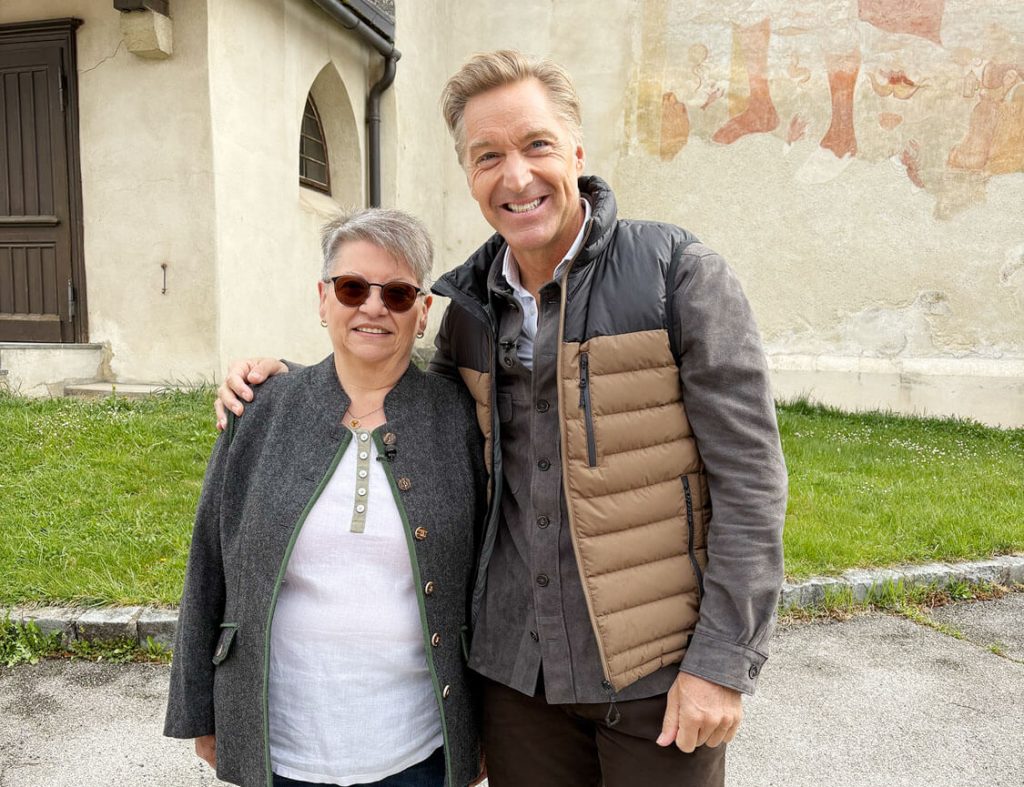 Zwei Personen stehen lächelnd vor einer Kirchenwand mit Wandmalerei und haben einander freundschaftlich den Arm umgelegt.