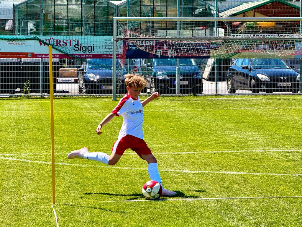 Das Dream-Team im Nachwuchs-Fußball 1 Ein junger Fußballspieler im rot-weißen Trikot führt einen Eckball aus. Mit konzentriertem Blick holt er zum Schuss aus, während der Ball auf der Ecklinie liegt und das Tor im Hintergrund zu sehen ist.